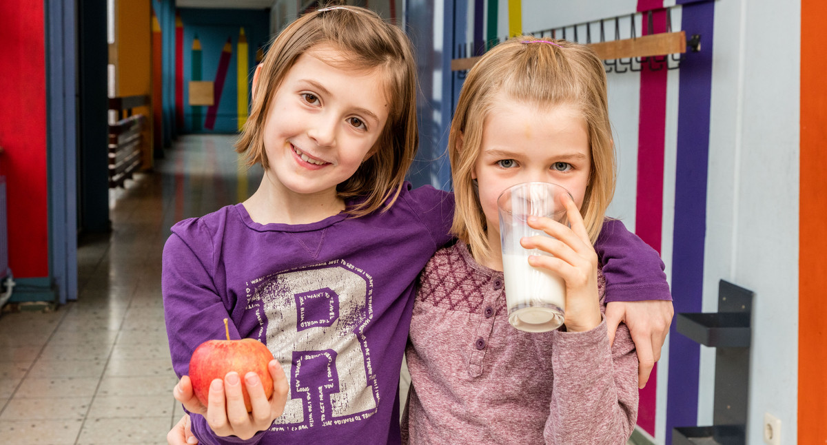 Mädchen im Schulflur mit Milchglas und Apfel.